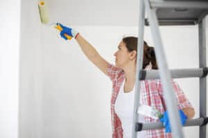 Young woman with paint roller and brush on ladder painting walls in her apartment.